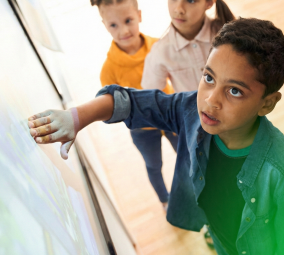 Young boy touching an interactive digital screen while two children watch behind him.